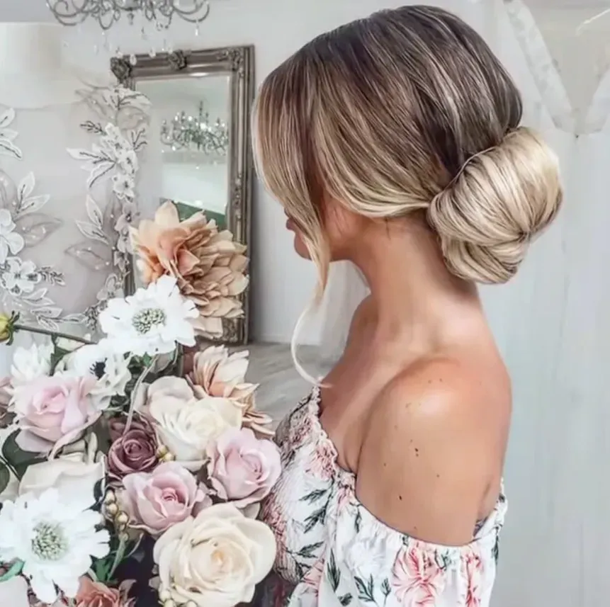 Textured low bridal bun with soft face-framing tendrils, beside an arrangement of flowers